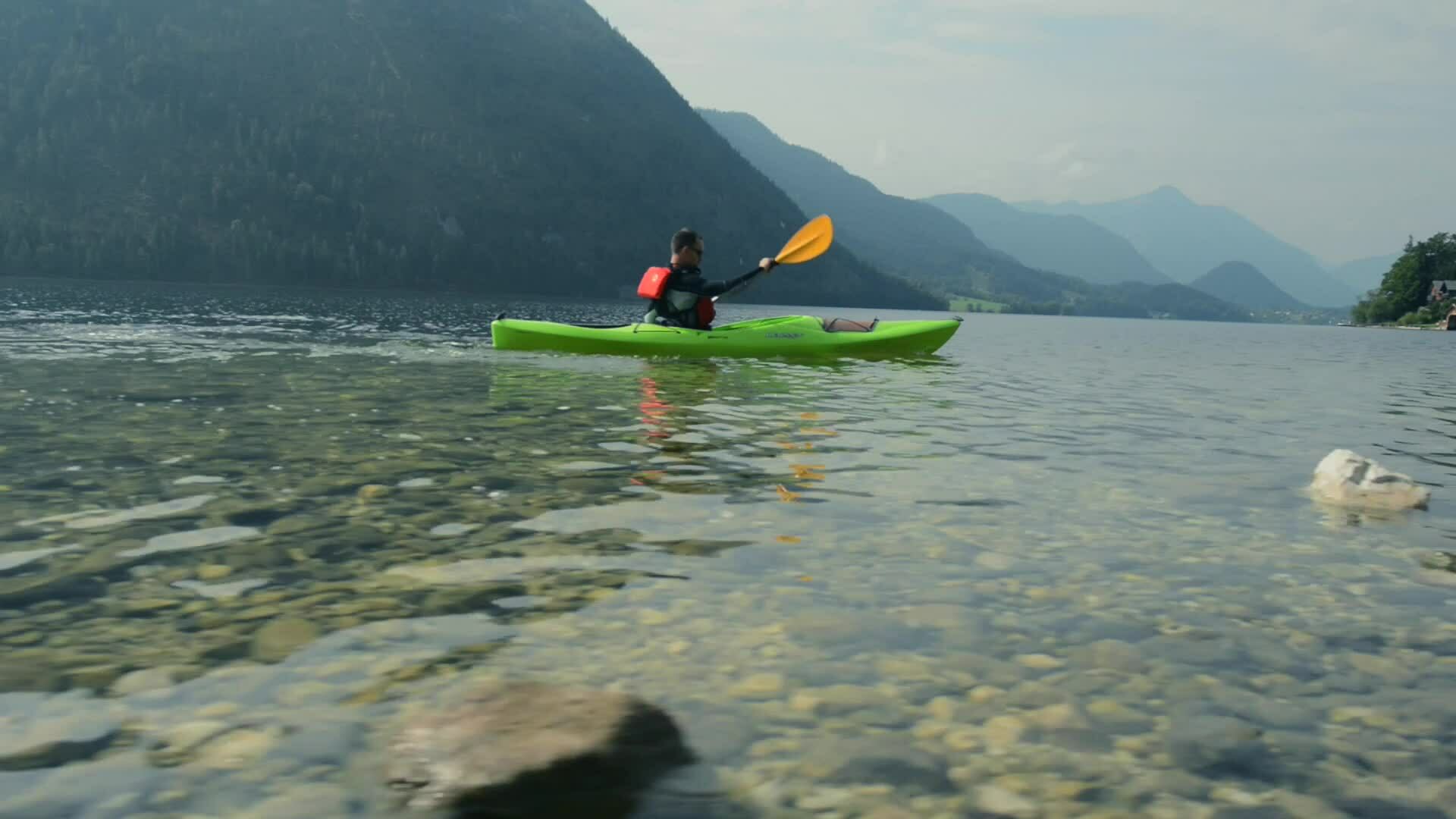 A kayaker in a green kayak paddling across a clear mountain lake with forested peaks in the background